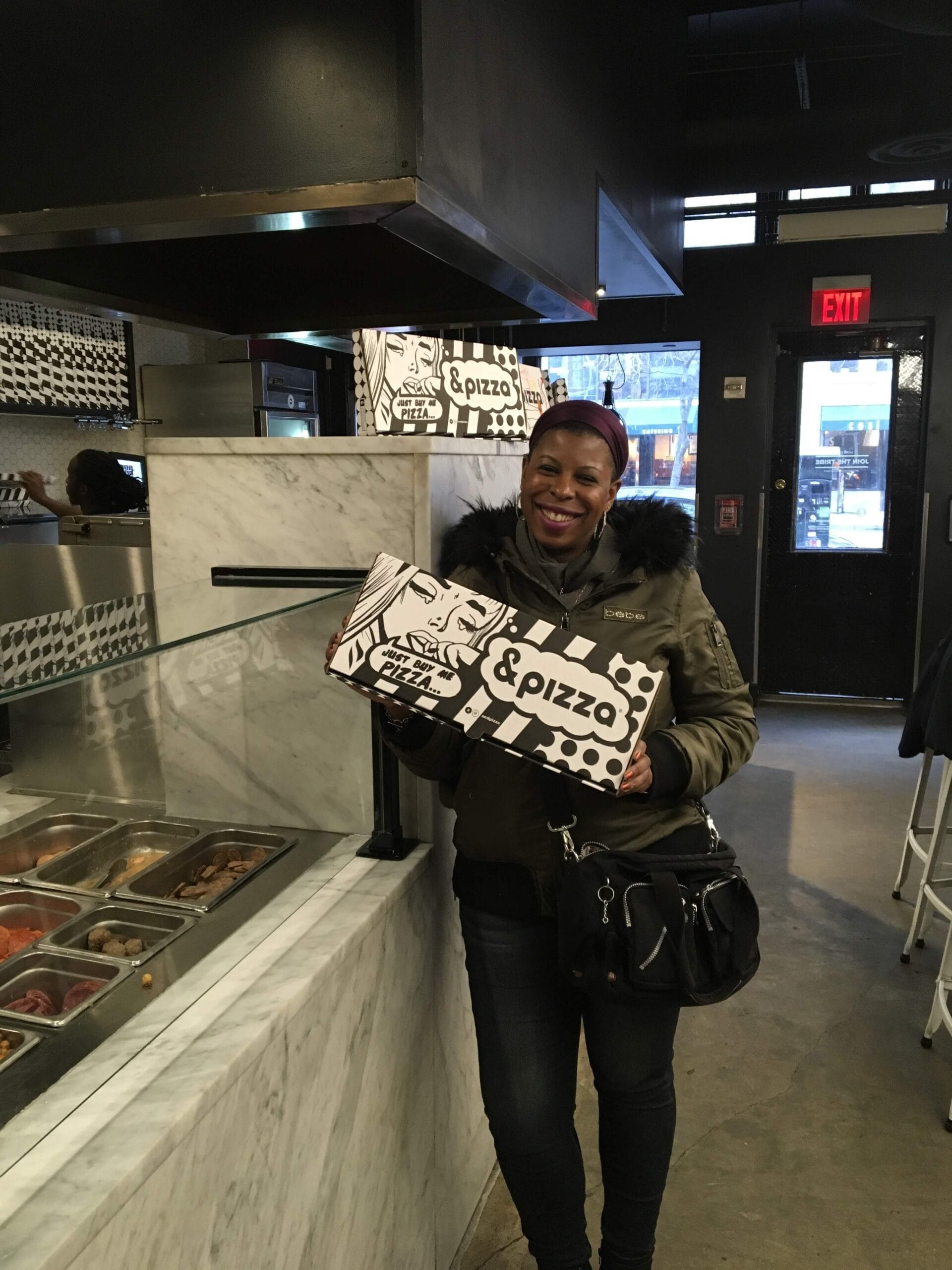 DG Speaks - Food - Review - Mercedes joyfully stands inside &pizza, cradling pizza boxes in her arms, ready to savor the gourmet delight.