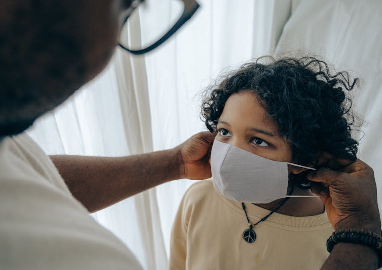 From above unrecognizable black man in healthcare wearing protective mask on face of ethnic kid standing in room near window closed with curtain