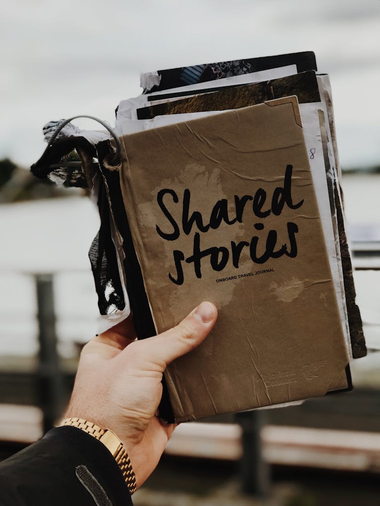 Hand holds a "Shared Stories" journal against a blurred outdoor backdrop, emphasizing creativity and personal storytelling.