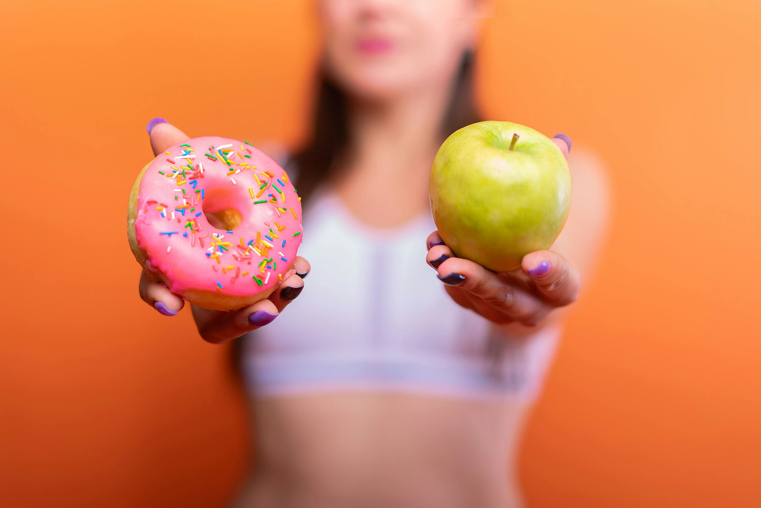 International No Diet Day. A woman holds a green apple and a pink donut with sprinkles against an orange background, emphasizing choice.