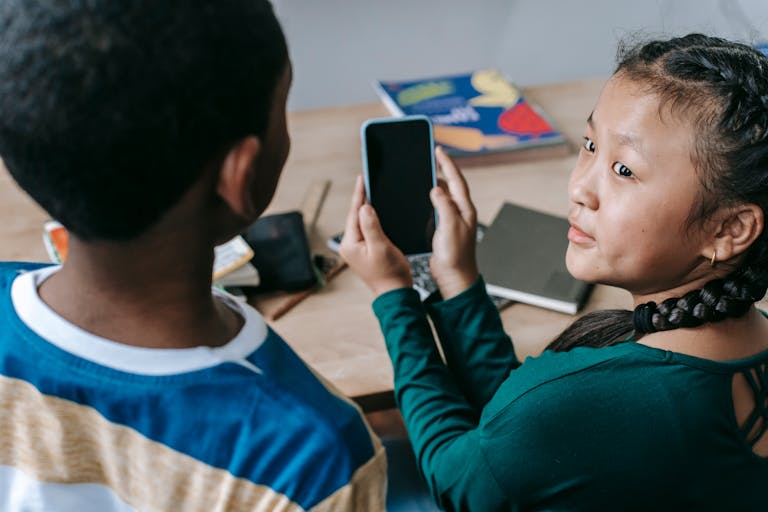 Mobile Learning in the Age of AI - From above of adorable little Asian girl showing mobile phone to unrecognizable African American boy during break in classroom