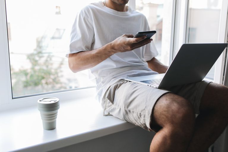 Travel Coffee and Comfort Essentials for Digital Nomads - Young man multitasking with smartphone and laptop while sitting by a window.