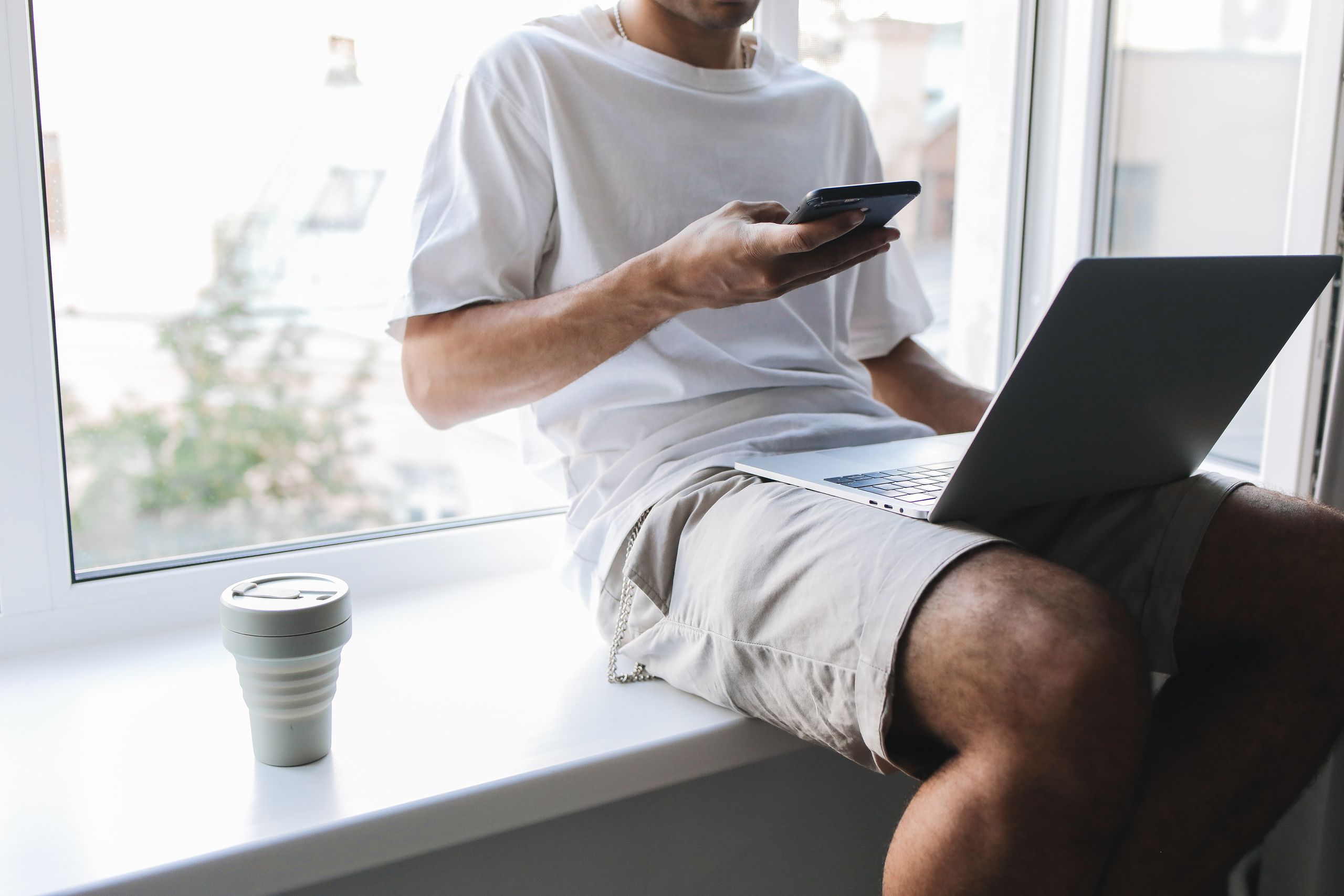 Travel Coffee and Comfort Essentials for Digital Nomads - Young man multitasking with smartphone and laptop while sitting by a window.