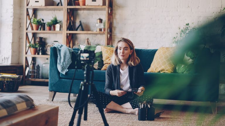 Woman filming vlog for Youtube at home with camera setup, surrounded by plants and cozy decor.