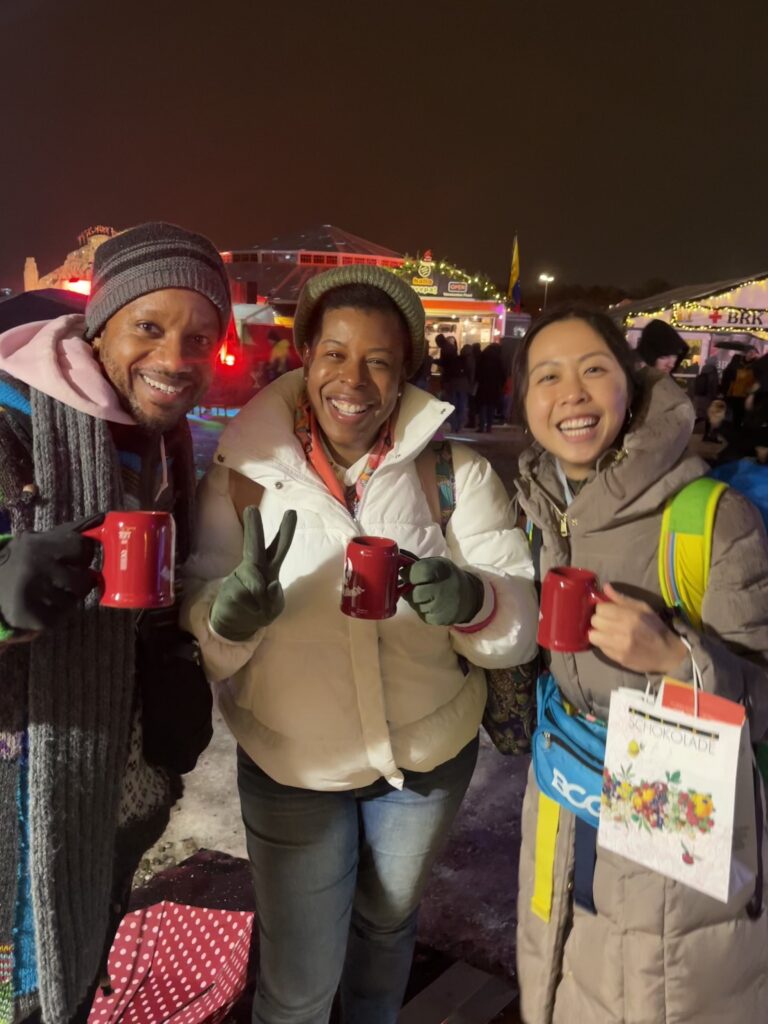 Why I Want Travel That Feels Like Belonging Practice - Three friends holding red mugs and smiling together at a festive German winter market at night