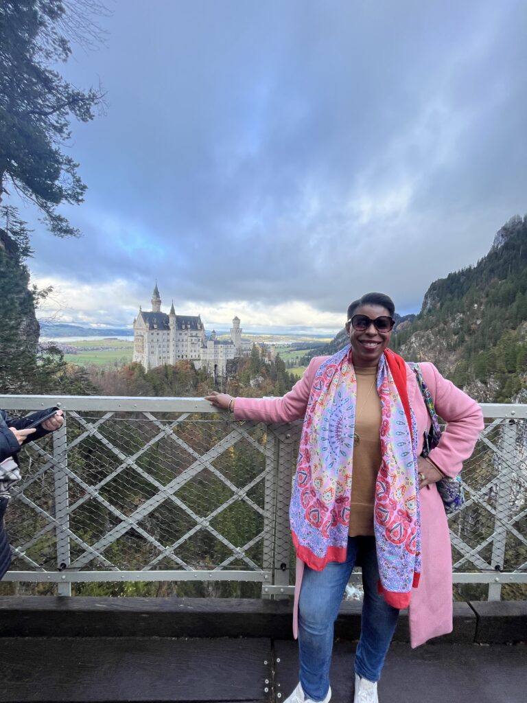 Black woman traveler wearing a pink coat with Neuschwanstein Castle behind her at Marienbrücke in Bavaria