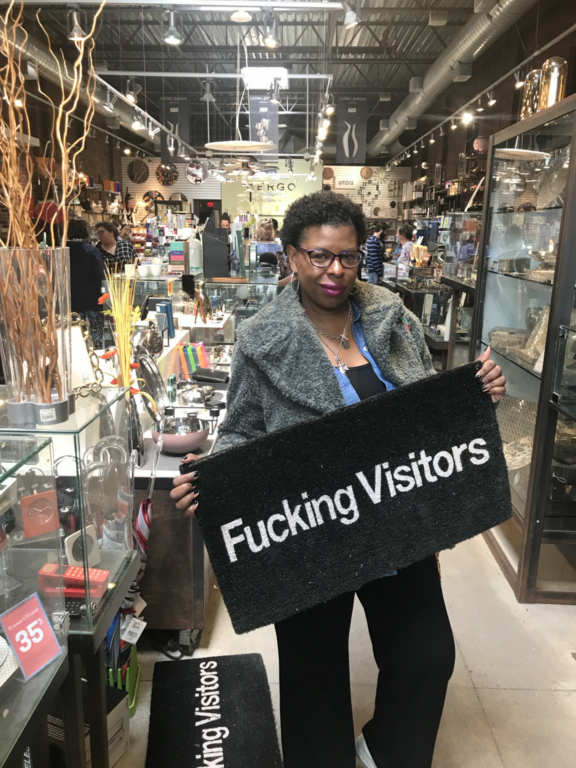 How to Handle Difficult Airbnb Guests Without Losing Your Cool - Mercedes Diane Griffin Forbes holding a humorous doormat while shopping, illustrating the importance of setting boundaries and handling difficult Airbnb guests.
