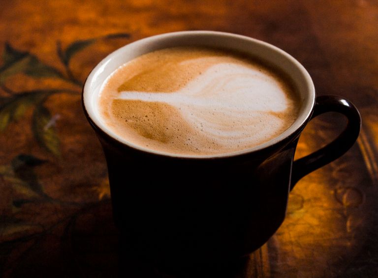 A close-up of a cappuccino in a black mug with latte art, against a warm, artistic background.