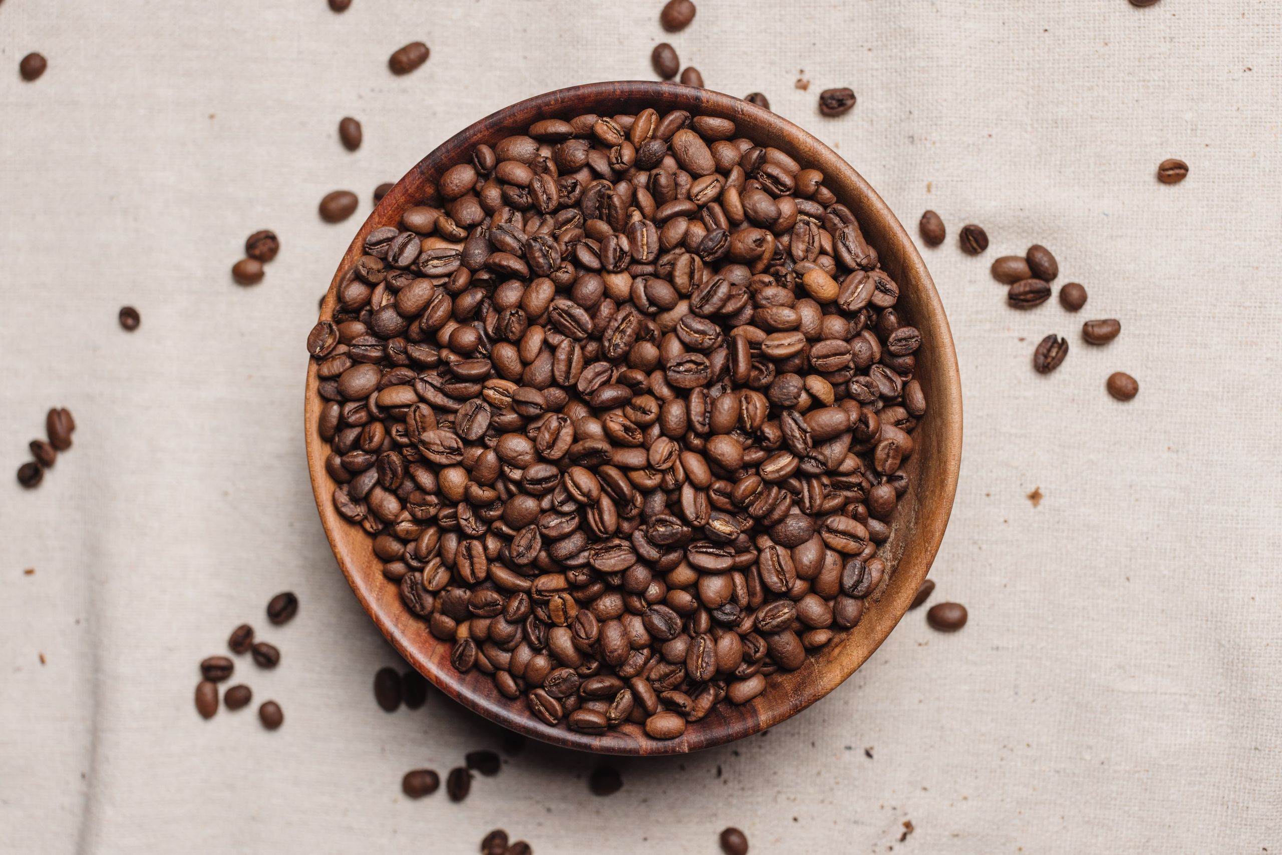 A detailed top view of fresh coffee beans in a wooden bowl on a cloth background.