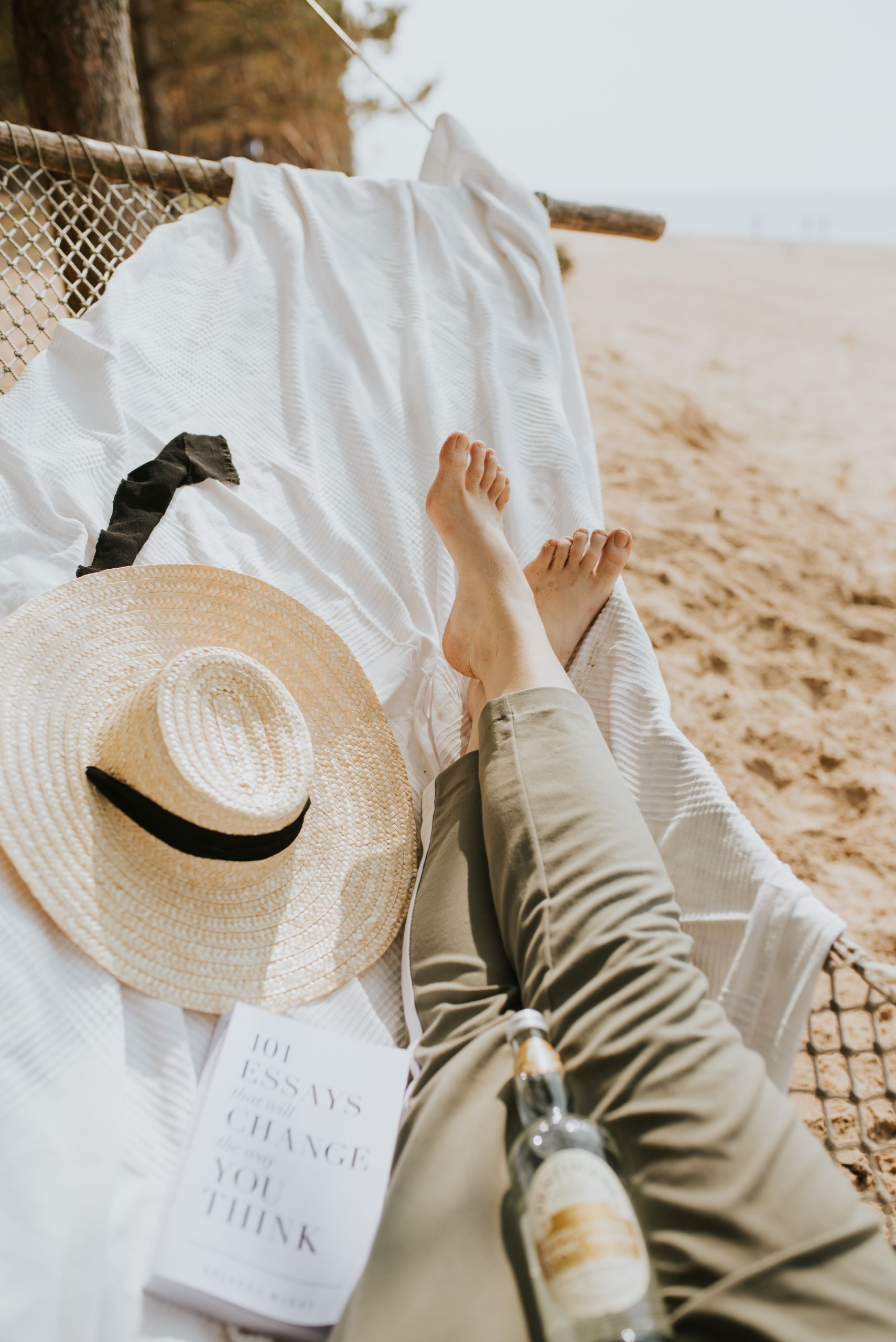 A person relaxes in a hammock on a sandy beach with a book and hat in view.
