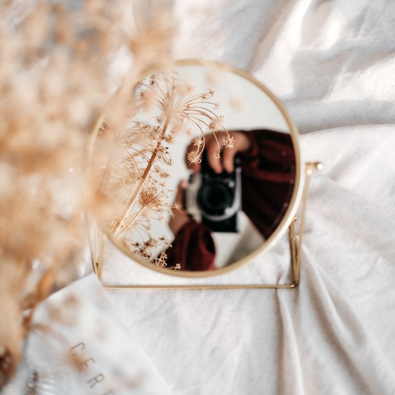 A round mirror reflecting a person holding a camera surrounded by delicate flowers. Soft and artistic composition.