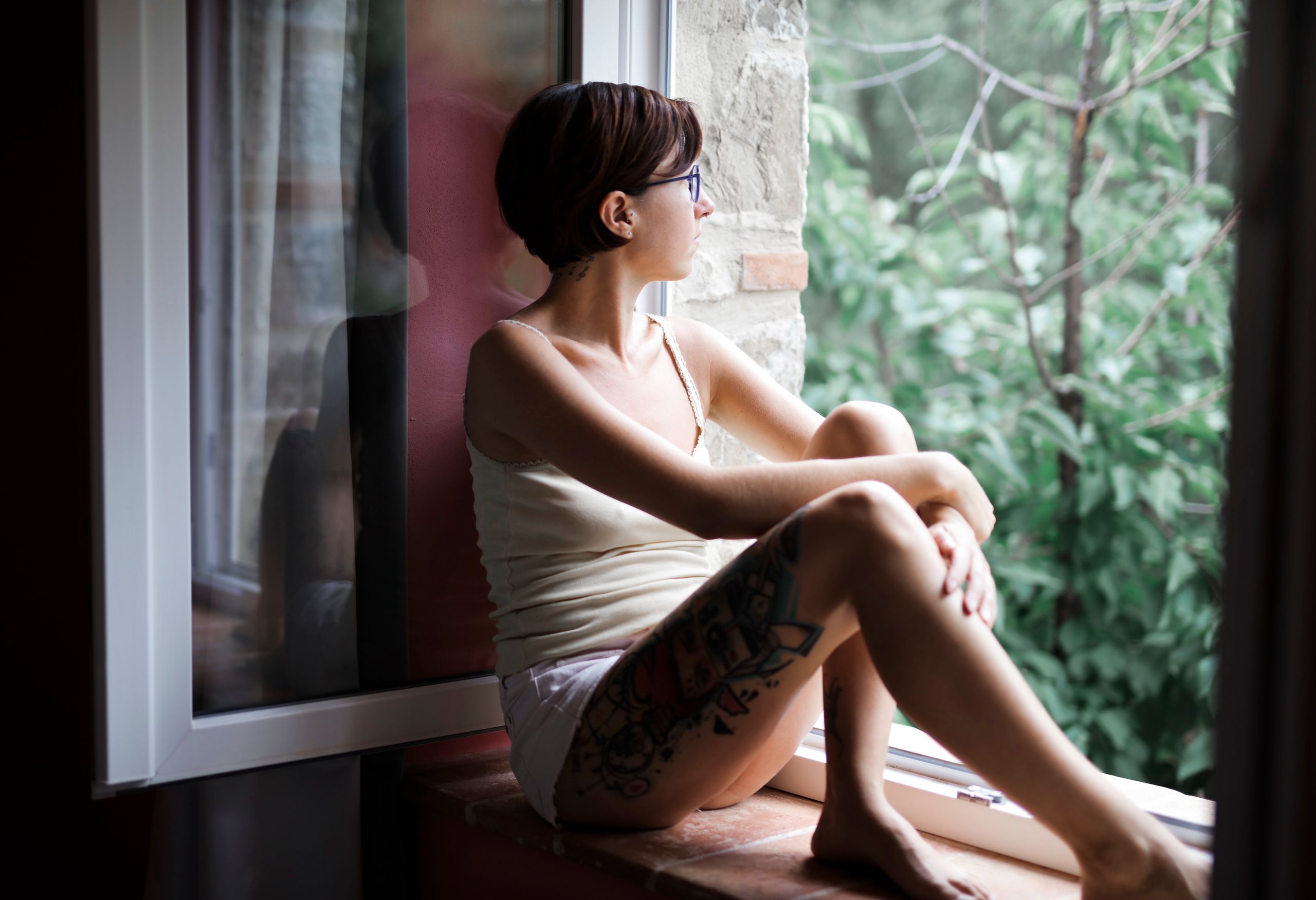 More Questions Than Answers - A tattooed woman with short hair sits by a window in deep thought, looking outside.