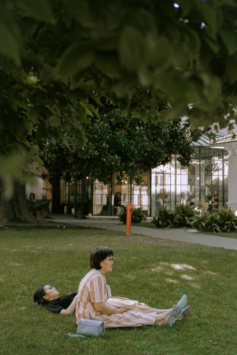 A Woman Resting in Public Is Still a Radical Sight - Two women relax on a green lawn under shade, enjoying a sunny day.