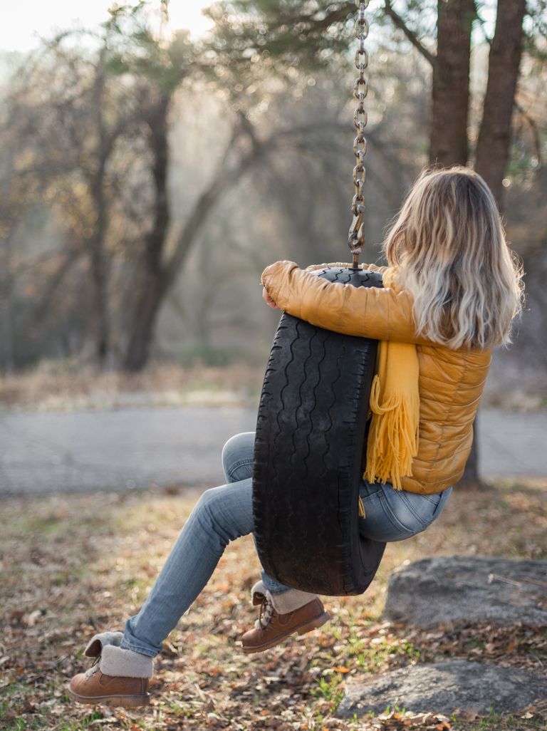 Braver Kind of Ease - Back view of anonymous lady in jacket and warm boots relaxing on tyre tree swing on quiet street on sunny autumn day