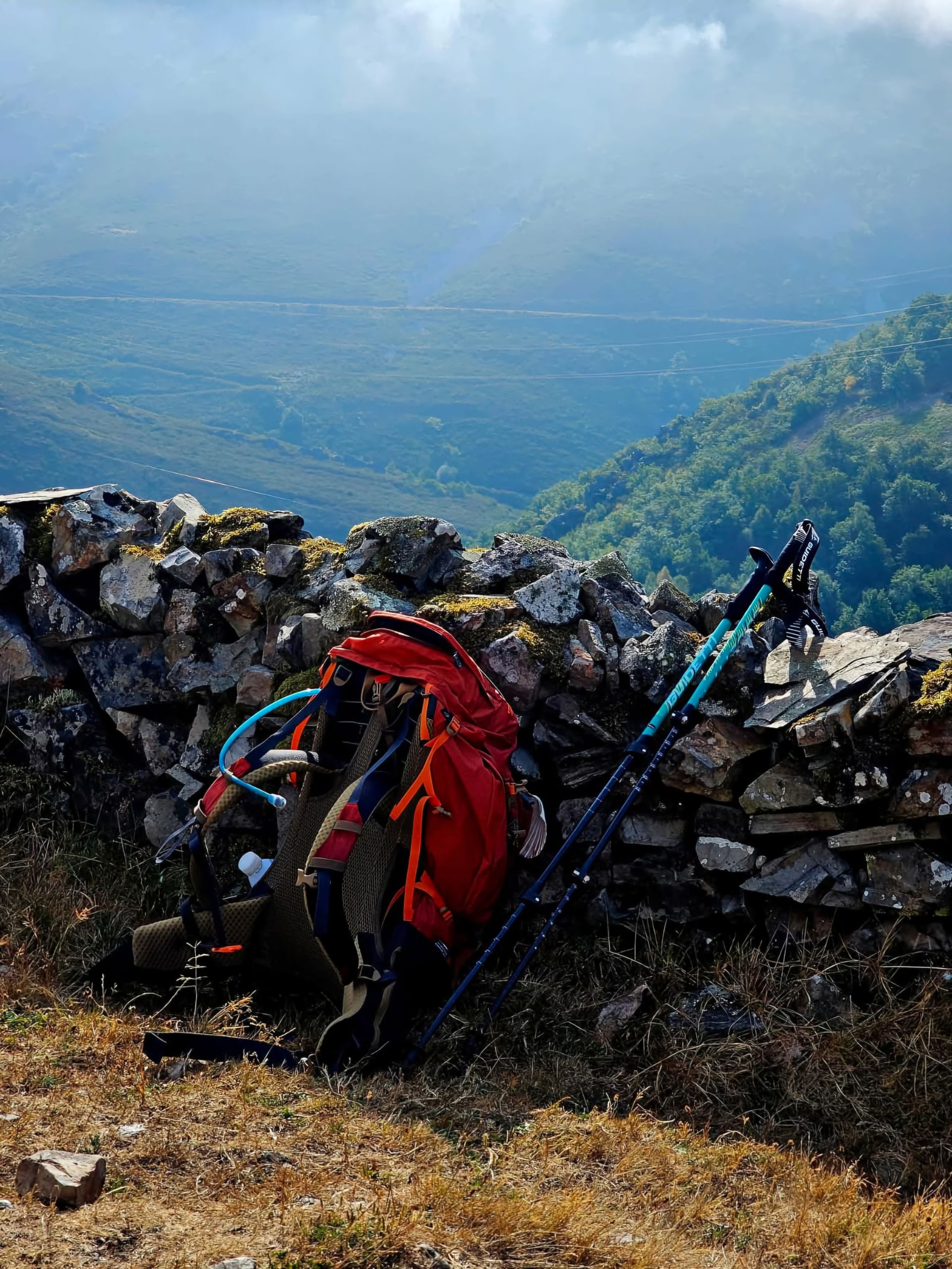 Camino Gear Guide: What Helped and What I Would Change - A red backpack with hiking poles against a stone wall overlooking a lush mountain landscape.