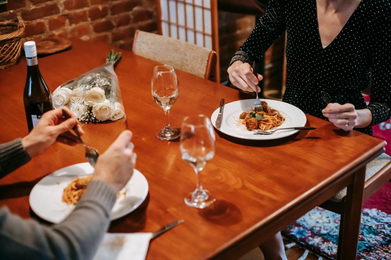 The Meal That Makes a Regular Day Feel Special - Crop faceless couple sitting in chairs at table while having dinner with pasta with meatballs on plates near cutlery and bottle with wine near glasses and flowers bouquet in room