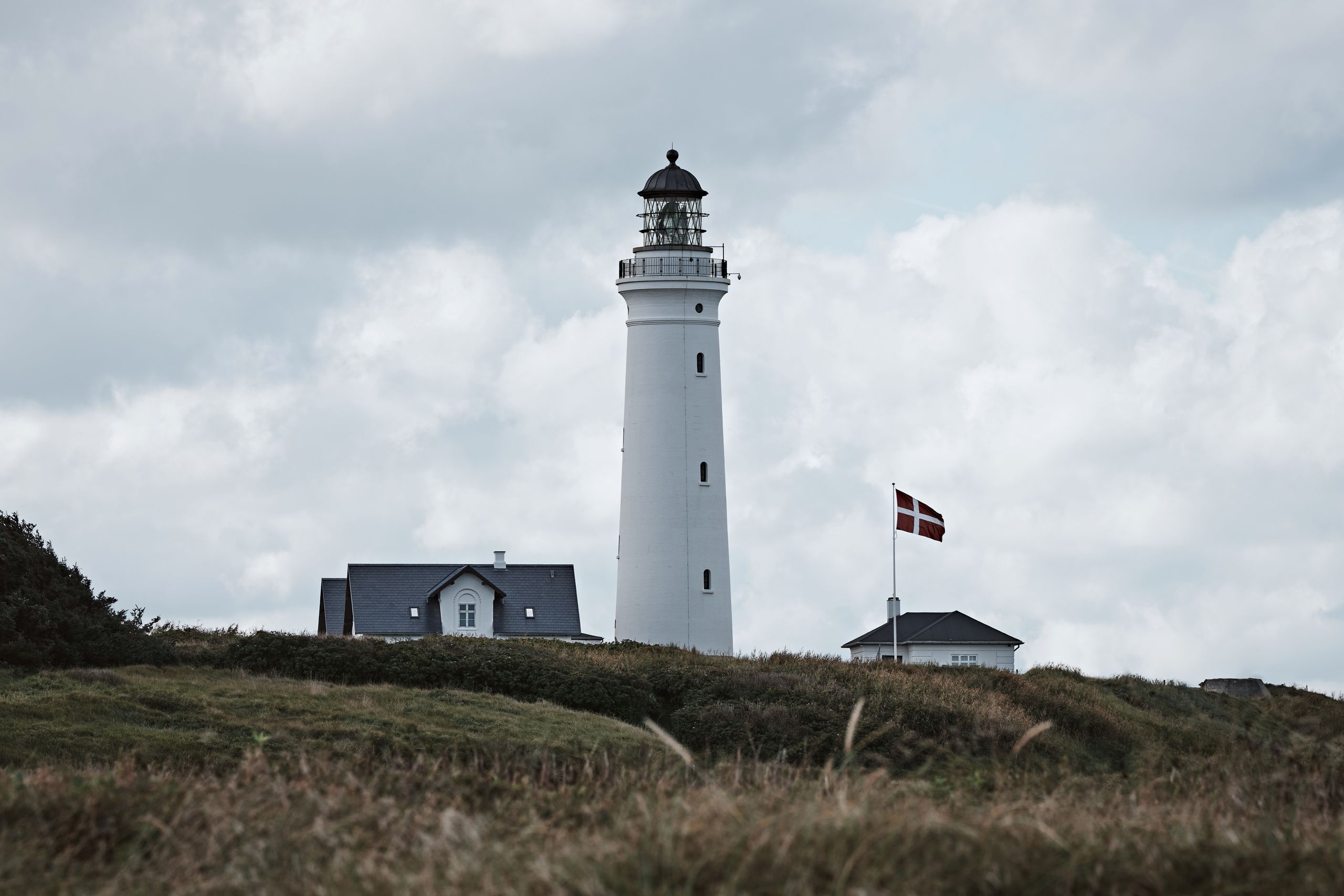Denmark Travel Guide - A scenic view of Hirtshals Lighthouse, Denmark, with a Danish flag, captured on a cloudy day.