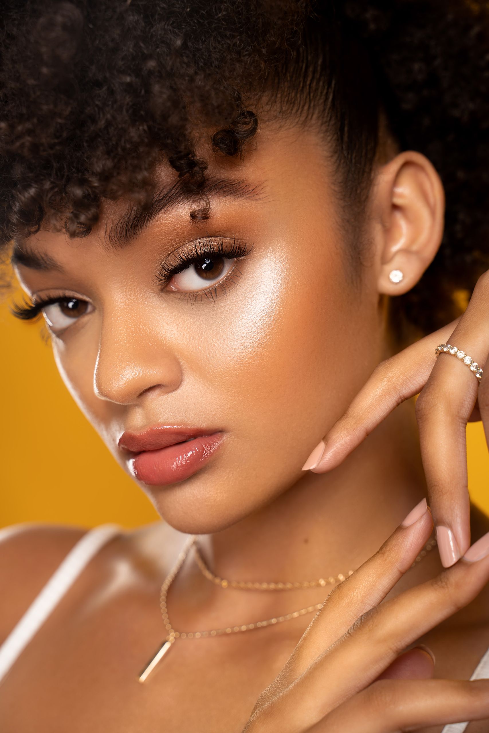 Glowing Skin Favorites- Close-up portrait of a young woman with curly hair, posing elegantly against a yellow background.