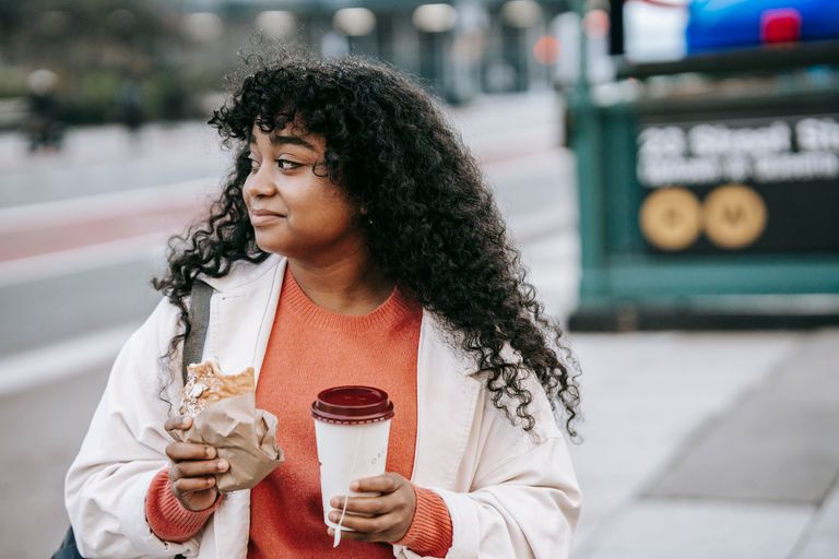 Hunger Is a Terrible Tour Guide - Glad adult African American female in casual outfit enjoying tasty sandwich and takeaway coffee while standing on city street and looking away