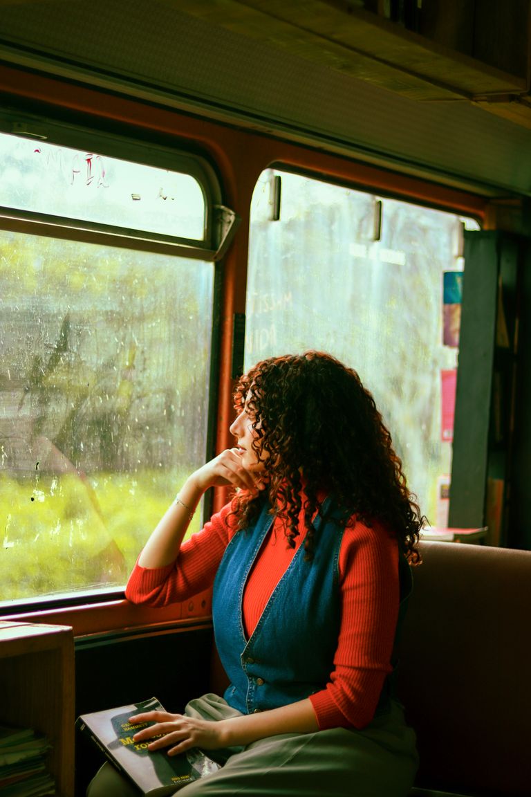 I Want a Life That Still Leaves Room for Wonder - Curly-haired woman looks out train window while holding a book, during a daytime journey.