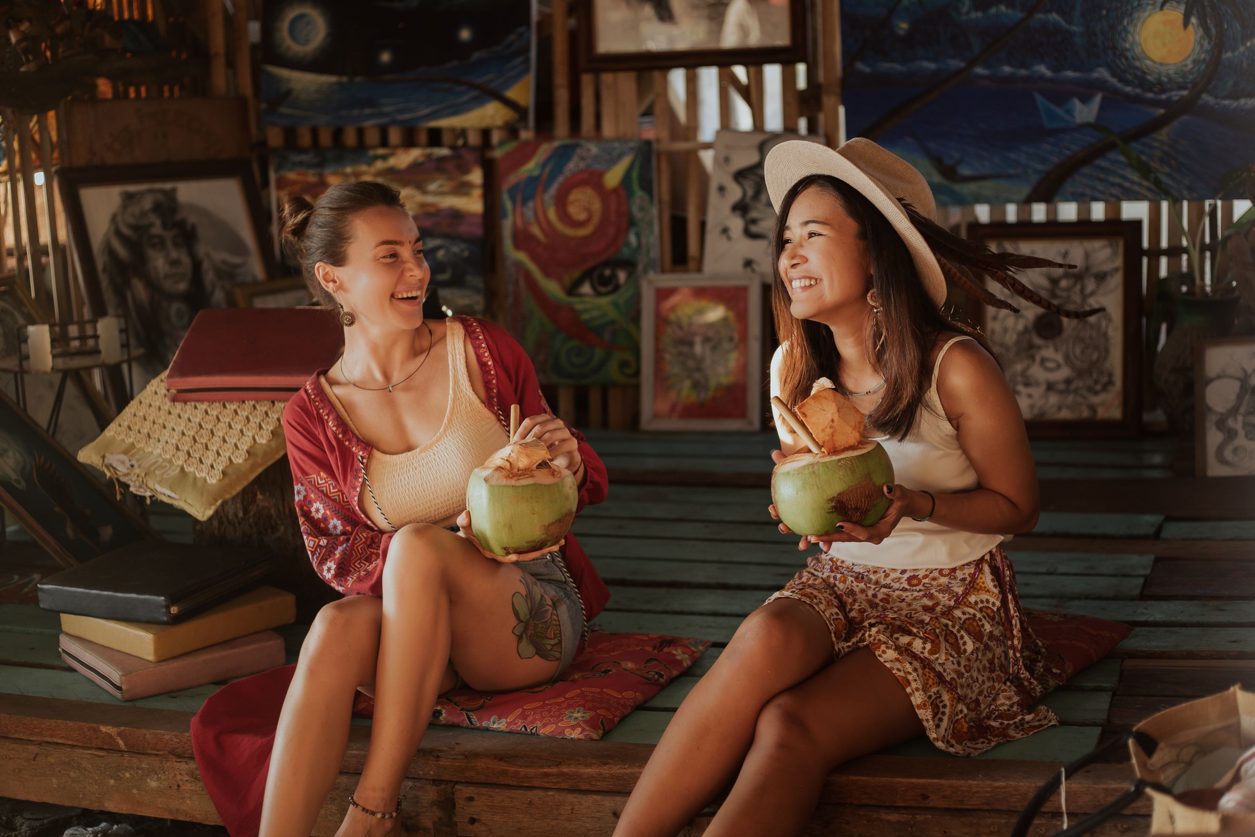 I Want Travel to Make Me More Useful - Two women laughing while enjoying refreshing coconut drinks in an artistic indoor setting.