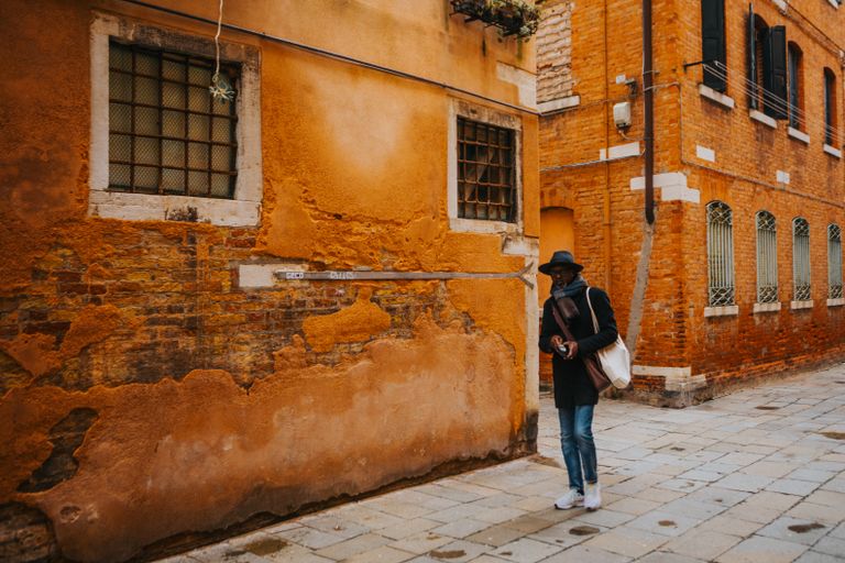 I Want Travel With Texture - A person in a hat explores a charming historic European alleyway with rustic brick buildings.
