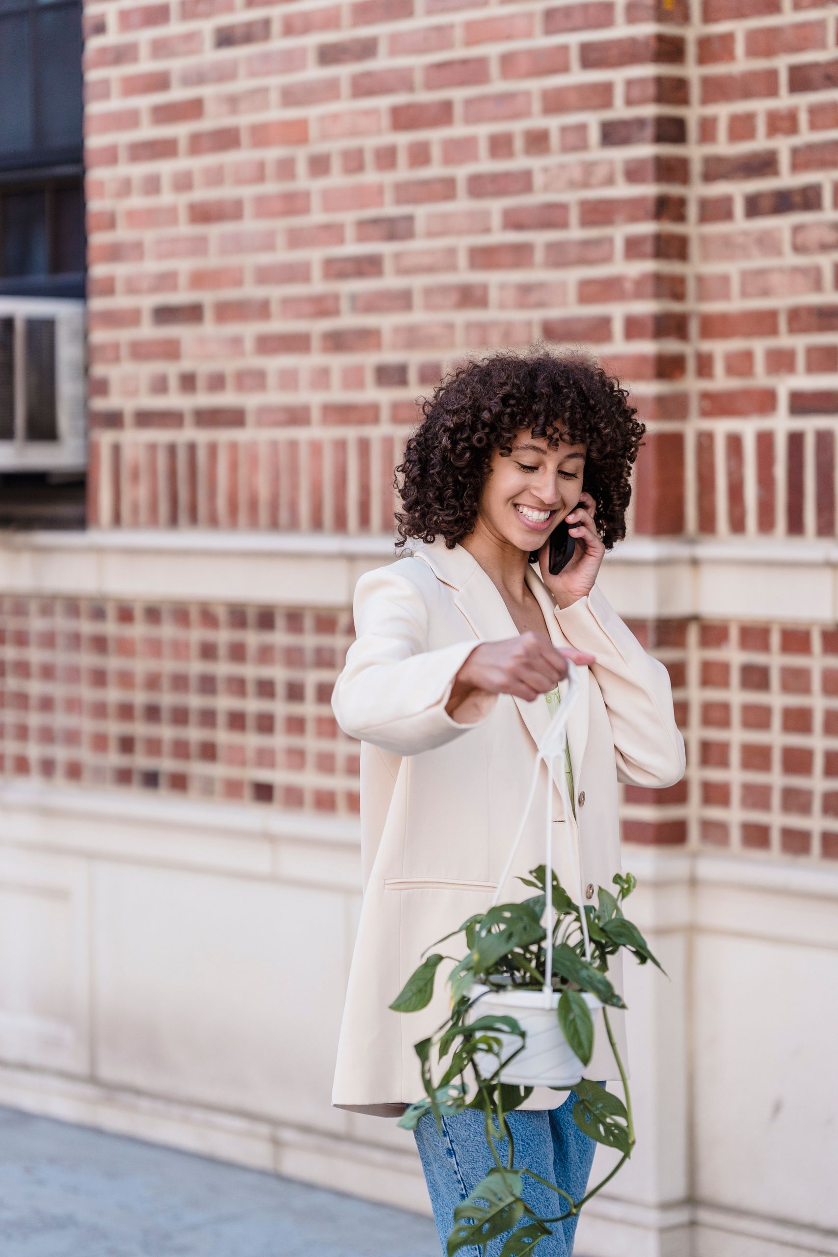 Learning to Ask for What I Need - Happy African American woman with smile talking on phone and carrying hanging flower pot while walking along tiled building wall