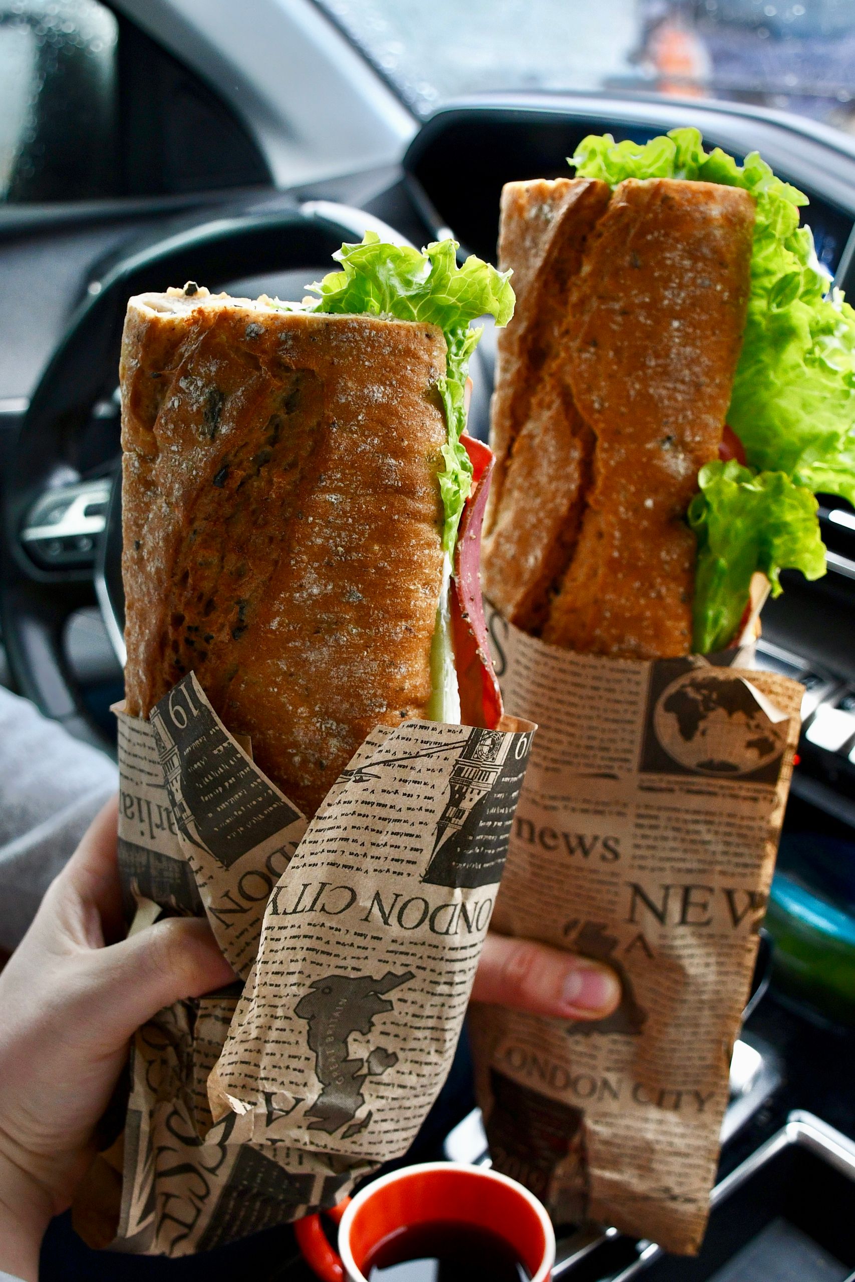 Love Food That Comes Wrapped in Paper - Two large sandwiches wrapped in newspaper held inside a car, with fresh lettuce and bread.