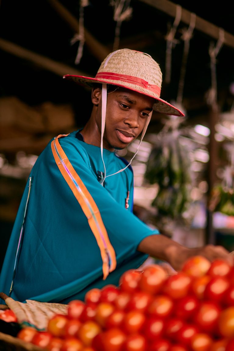 Market Tomato - A young man in a colorful hat examines ripe tomatoes at a bustling outdoor market.