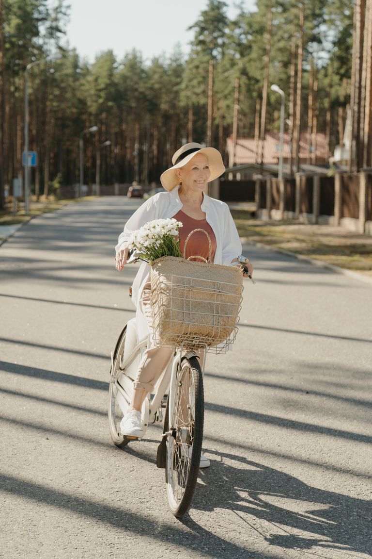 Pace Than Productivity -An elderly woman rides a bicycle outdoors, wearing a sun hat and basking in the sunlight.