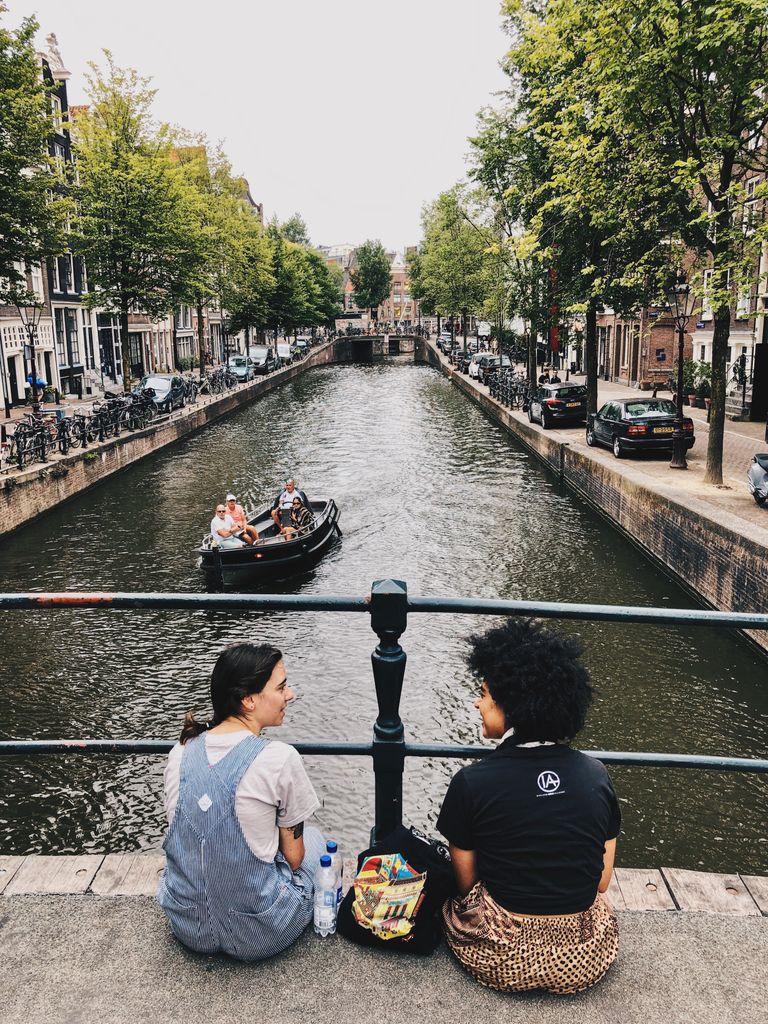 Slow Travel Helps a Place Become Real - Two women sitting on an Amsterdam bridge, enjoying a view of the canal and passing boat.