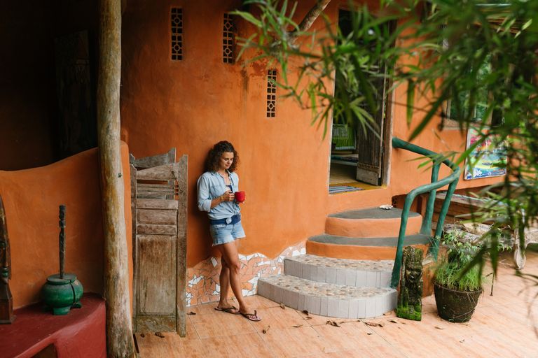 Slow Travel Helps Me See A Place Clearly - Full body brunette wearing denim clothes with cup of hot drink leaning on rural cottage wall while visiting exotic tropical country