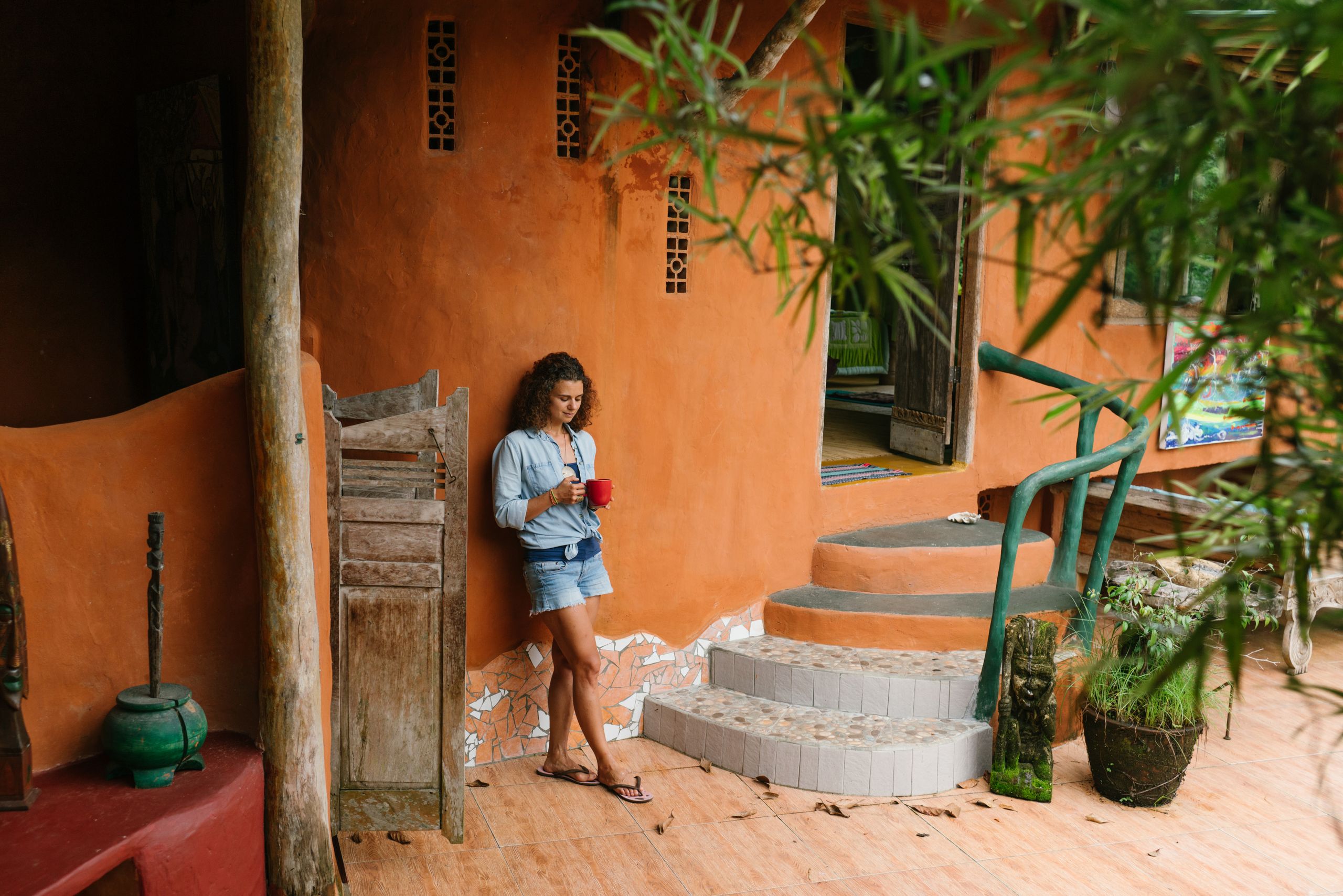 Slow Travel Helps Me See A Place Clearly - Full body brunette wearing denim clothes with cup of hot drink leaning on rural cottage wall while visiting exotic tropical country