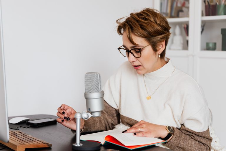 Telling Her Story - Female narrator in glasses reading out loud from book while sitting at desk with microphone and recording audiobook in cozy study