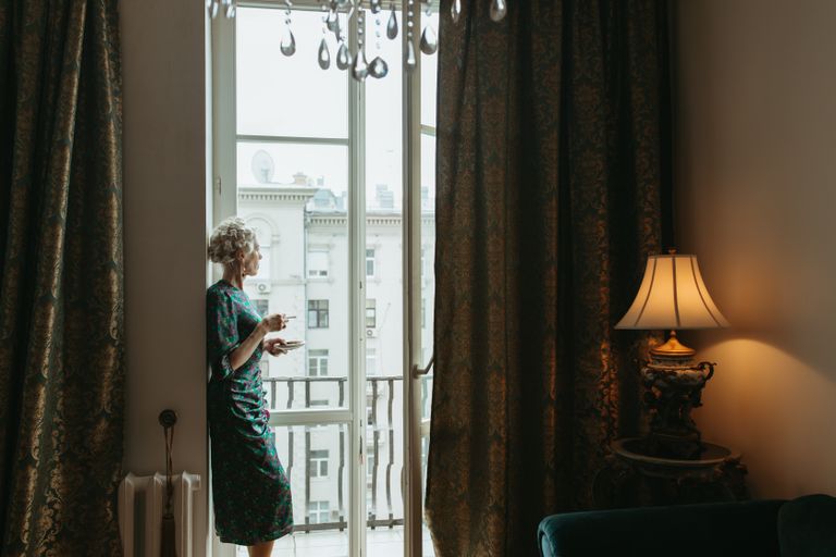 The Quiet Desire Beneath the Journey - A woman stands by a glass door in an elegantly decorated room with curtains and a lamp.