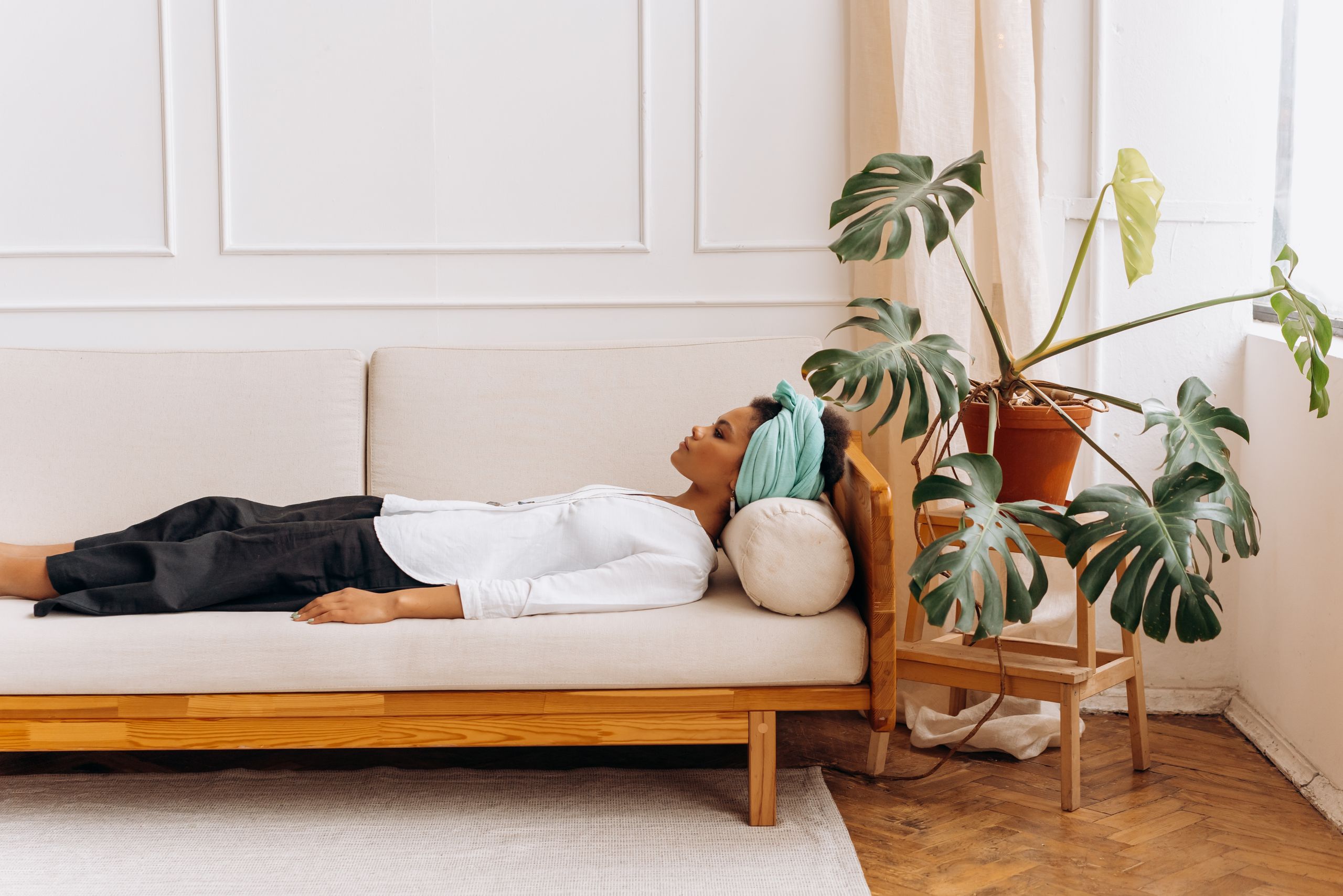 The Quiet Power of Rest for Women Who Lead - A woman relaxes on a sofa, wearing a headscarf, indoors with a large plant nearby.