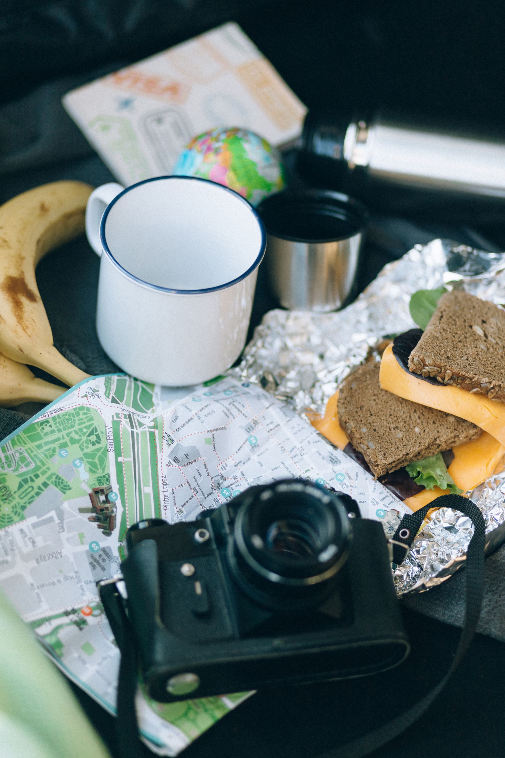 The Road Trip Snack Bag Is Serious Business - A flat lay of travel essentials including a map, camera, sandwich, and banana for a journey.