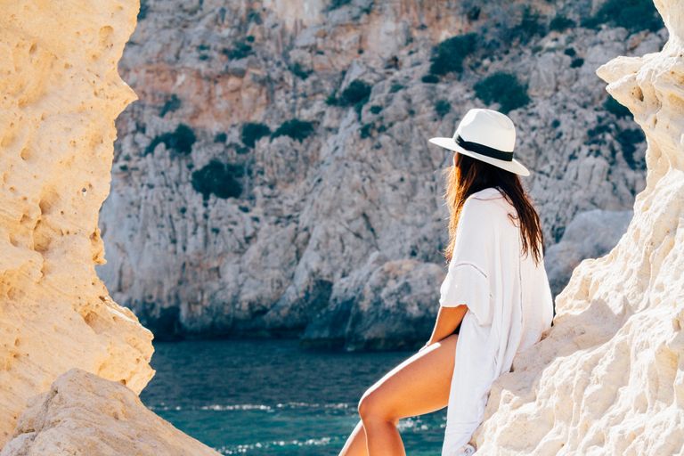 The Sun Gets a Vote: How I Travel Differently in the Heat - A woman in a hat sits on a rocky beach in summer, enjoying the sea view.