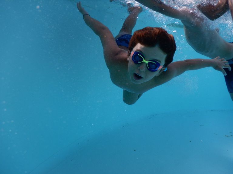 WATERINJOY Panoramic View Swim Goggles, 100% UV Protected with Siamese Ear Plugs - Young boy swimming underwater in a pool with goggles in bright summer light.