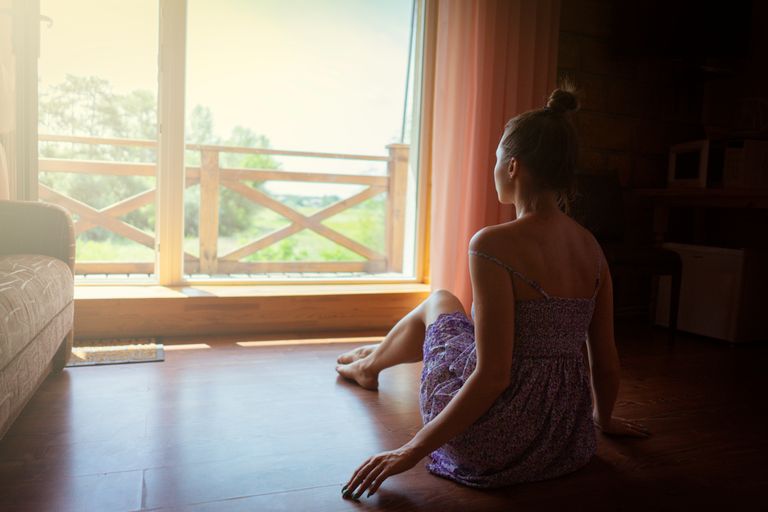 What a Quiet Room Can Give Back - A woman sits inside a sunlit room looking out a large window, enjoying nature.