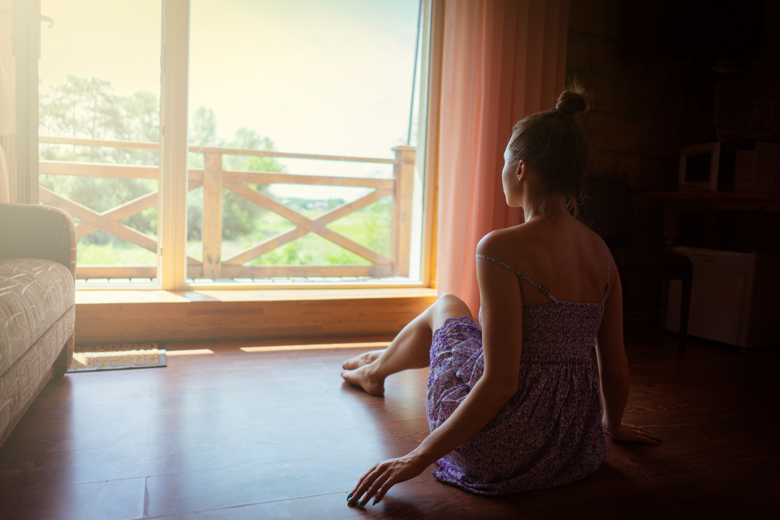 What a Quiet Room Can Give Back - A woman sits inside a sunlit room looking out a large window, enjoying nature.