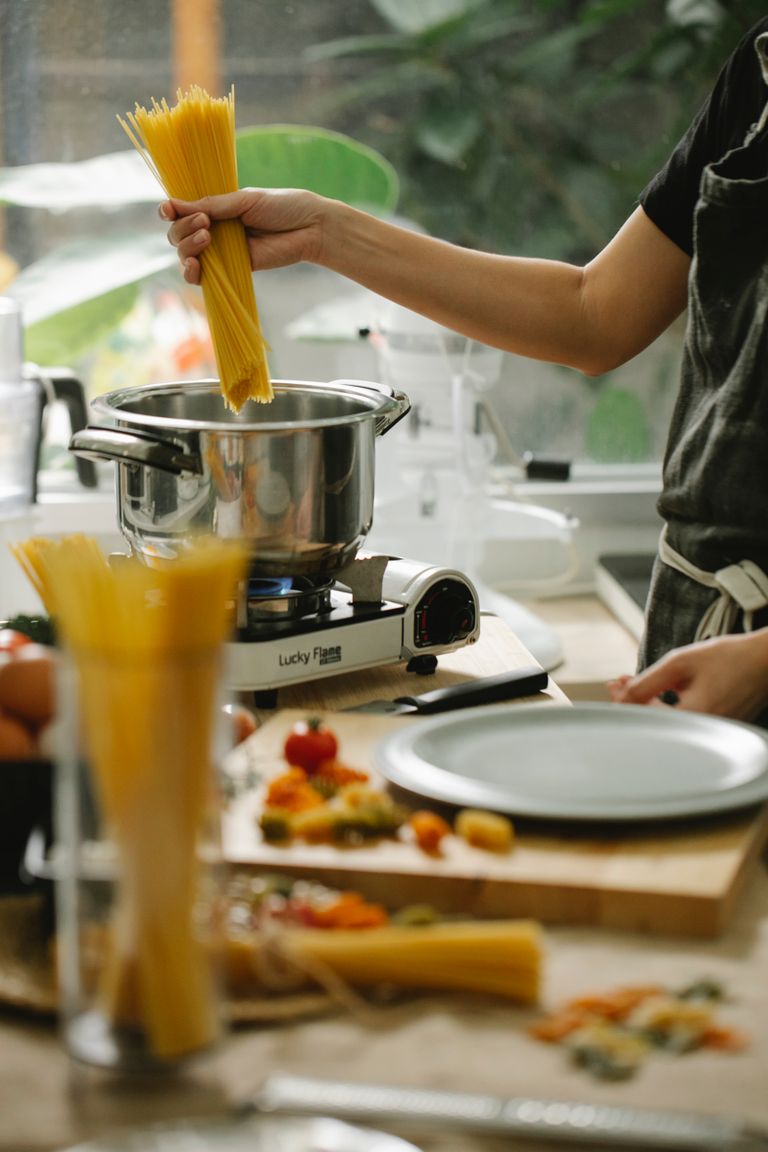 What Home Cooking Teaches About Survival -Crop unrecognizable female chef in apron putting raw spaghetti into saucepan on stove while cooking in contemporary light kitchen