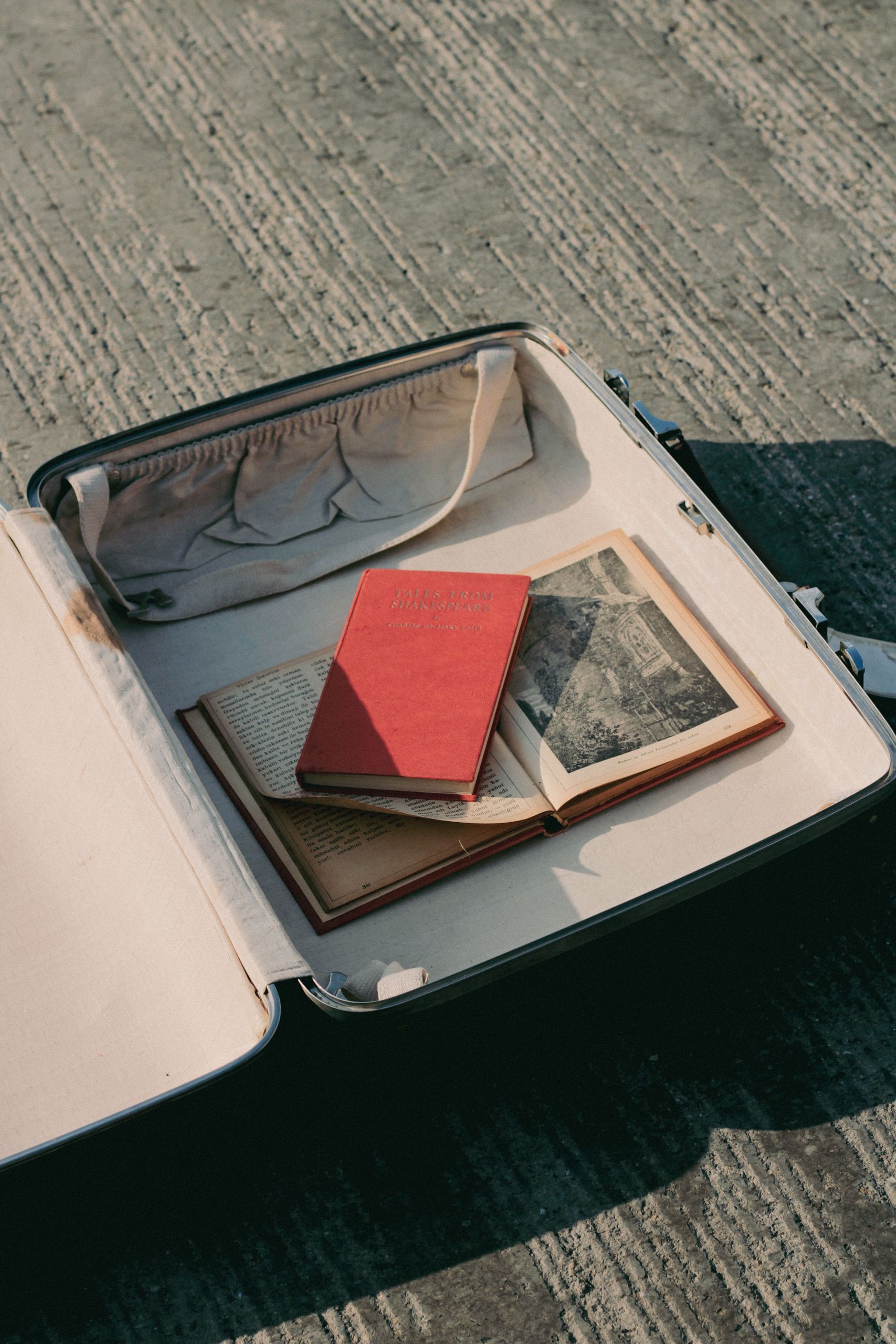 What I Am Carrying Into the Next Chapter - An open suitcase filled with vintage books on a textured concrete floor, evoking travel memories.