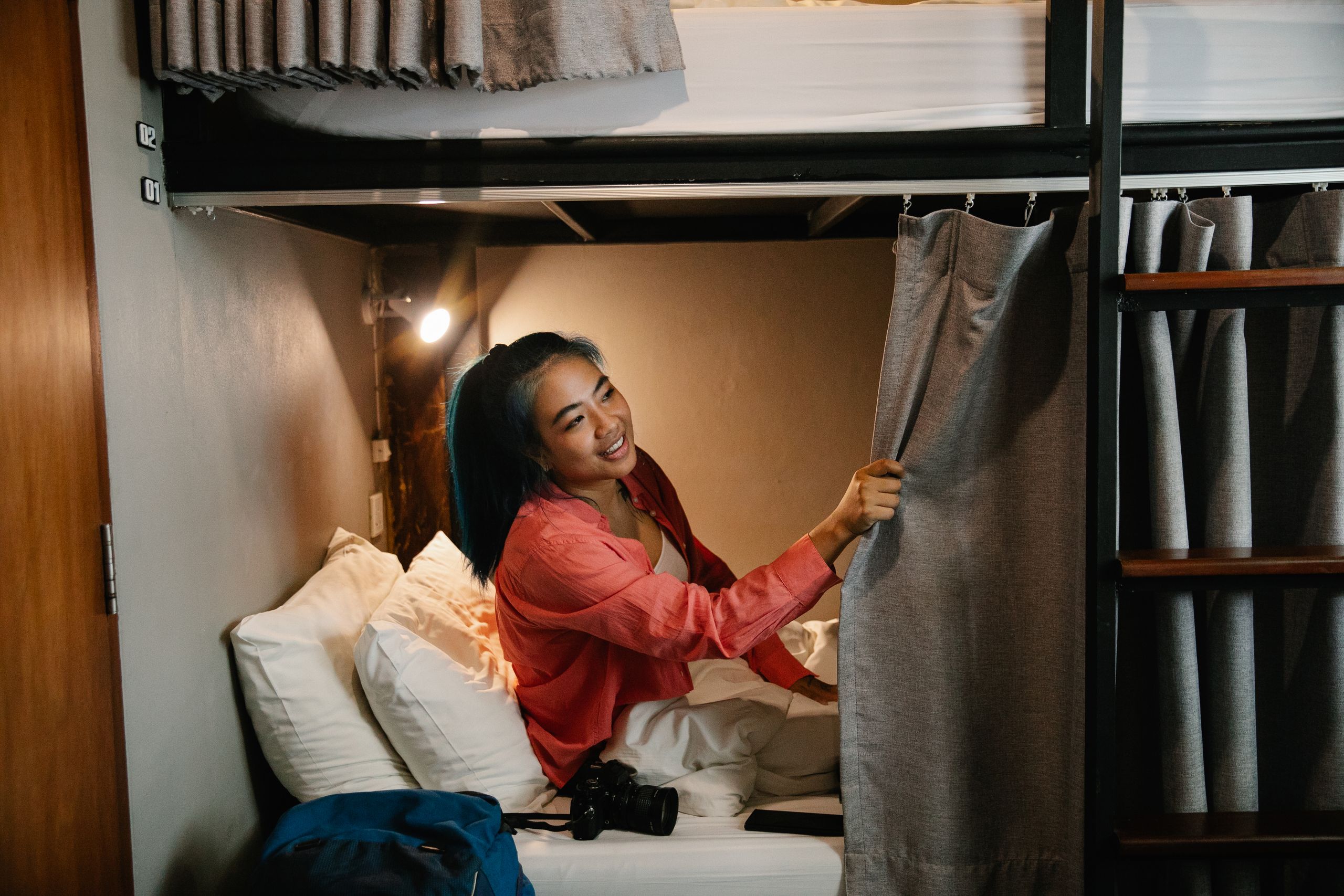 What It Means to Welcome a Woman Traveling Alone - Side view of young smiling ethnic female resting in bed under white blanket with camera and phone and looking away holding curtain in train