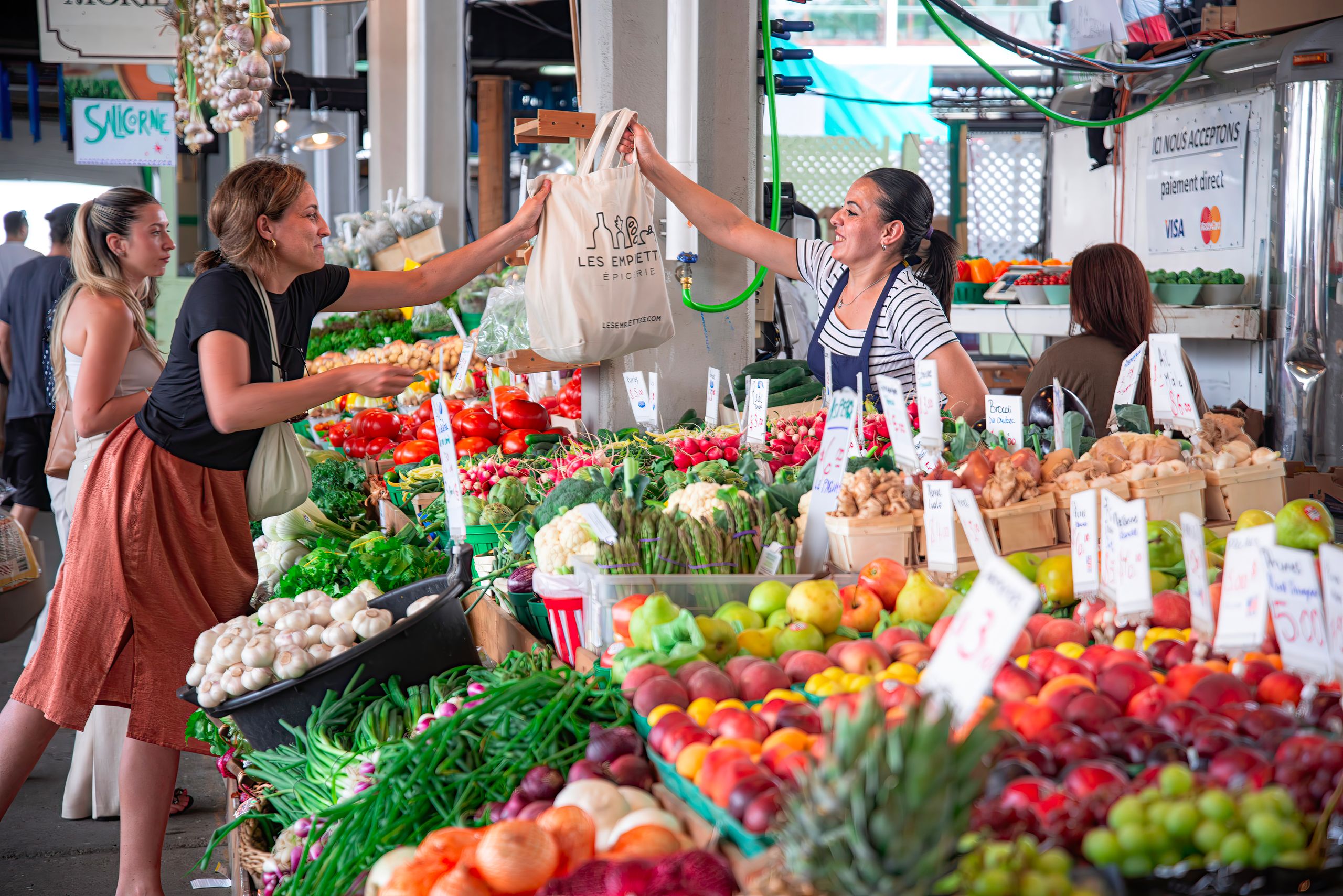 What Markets Teach Me About Community - Women shopping at a colorful outdoor market stall filled with fresh fruits and vegetables. Smiles and interaction.