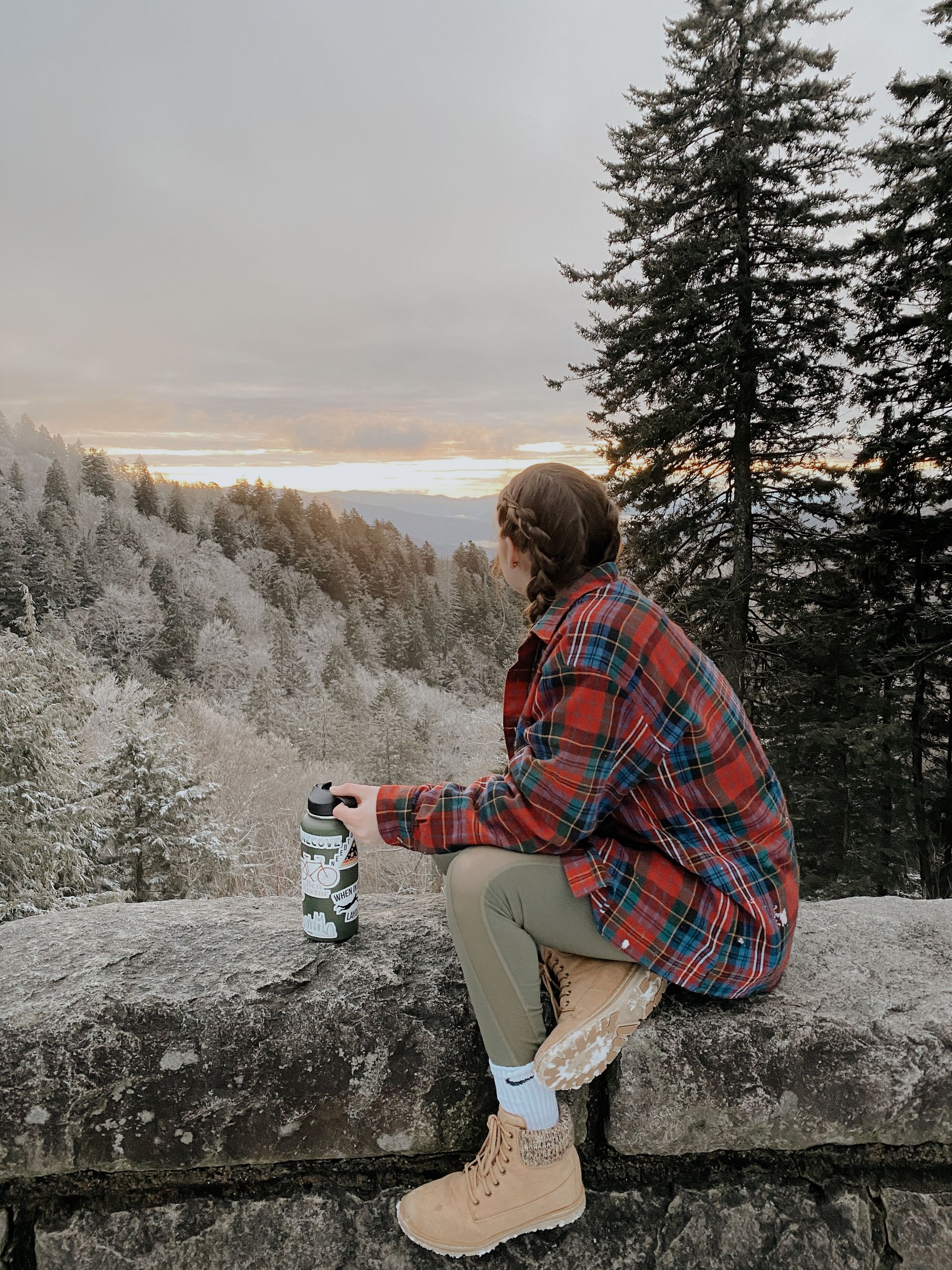 What This Year Taught Me About Enough - A woman in a plaid shirt sits on a rock wall, admiring a snowy mountain vista at sunset.