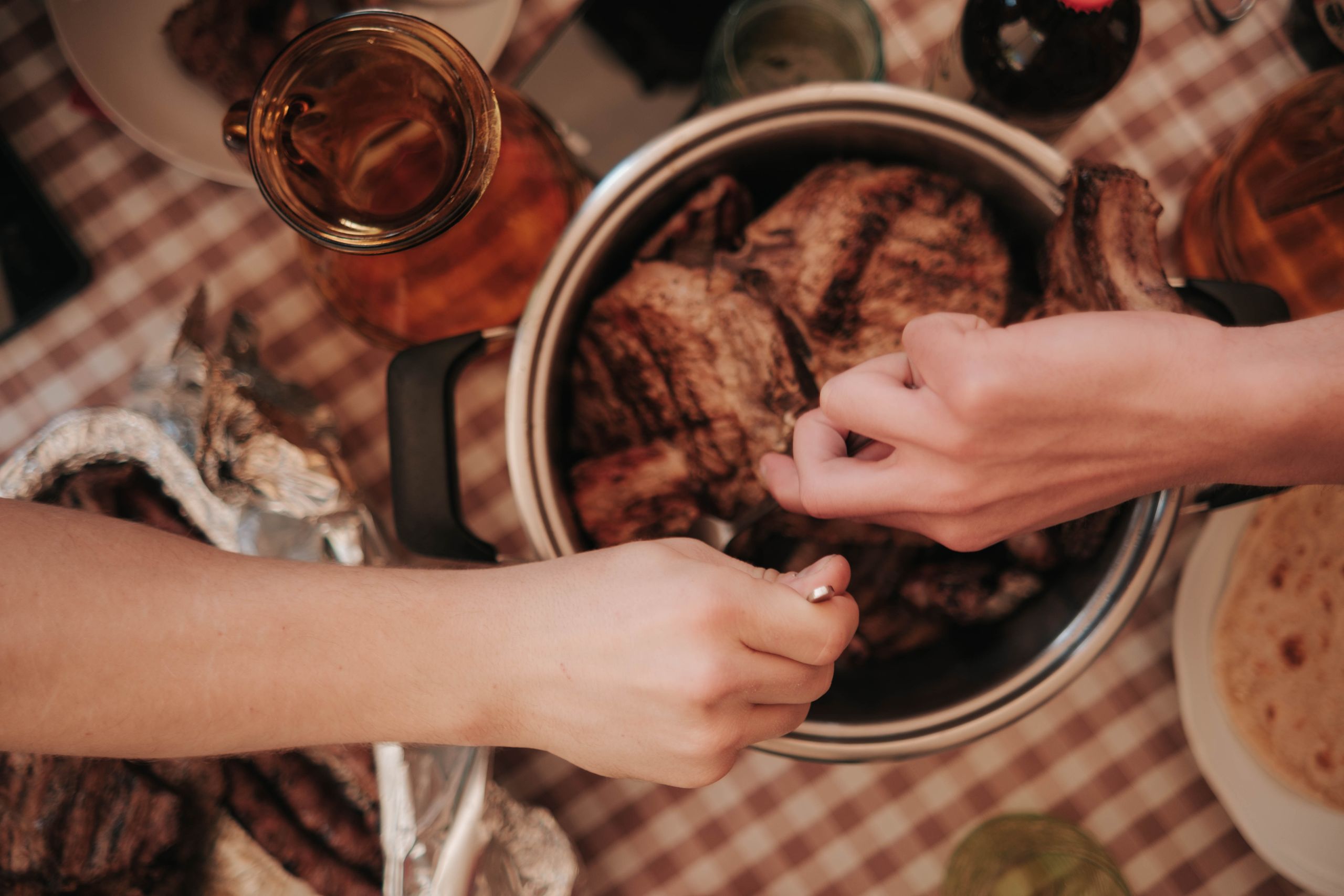 What We Learn When Everyone Eats From the Same Pot - Top-down view of hands serving roasted meat from a pot on a dining table.