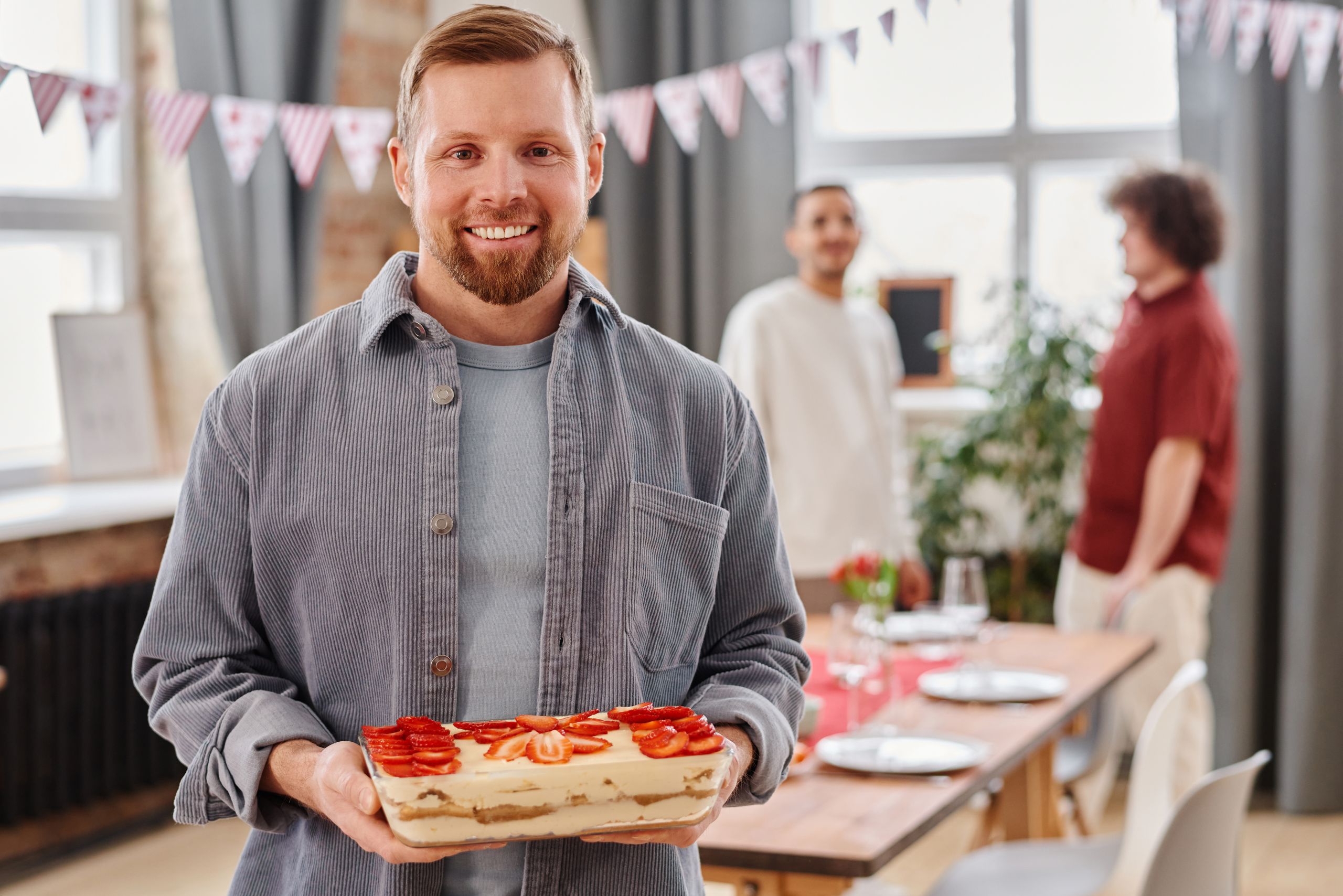 Why Bringing Extra Food Feels Like a Form of Welcome - Smiling man holding a strawberry cake at an indoor party with friends in the background.