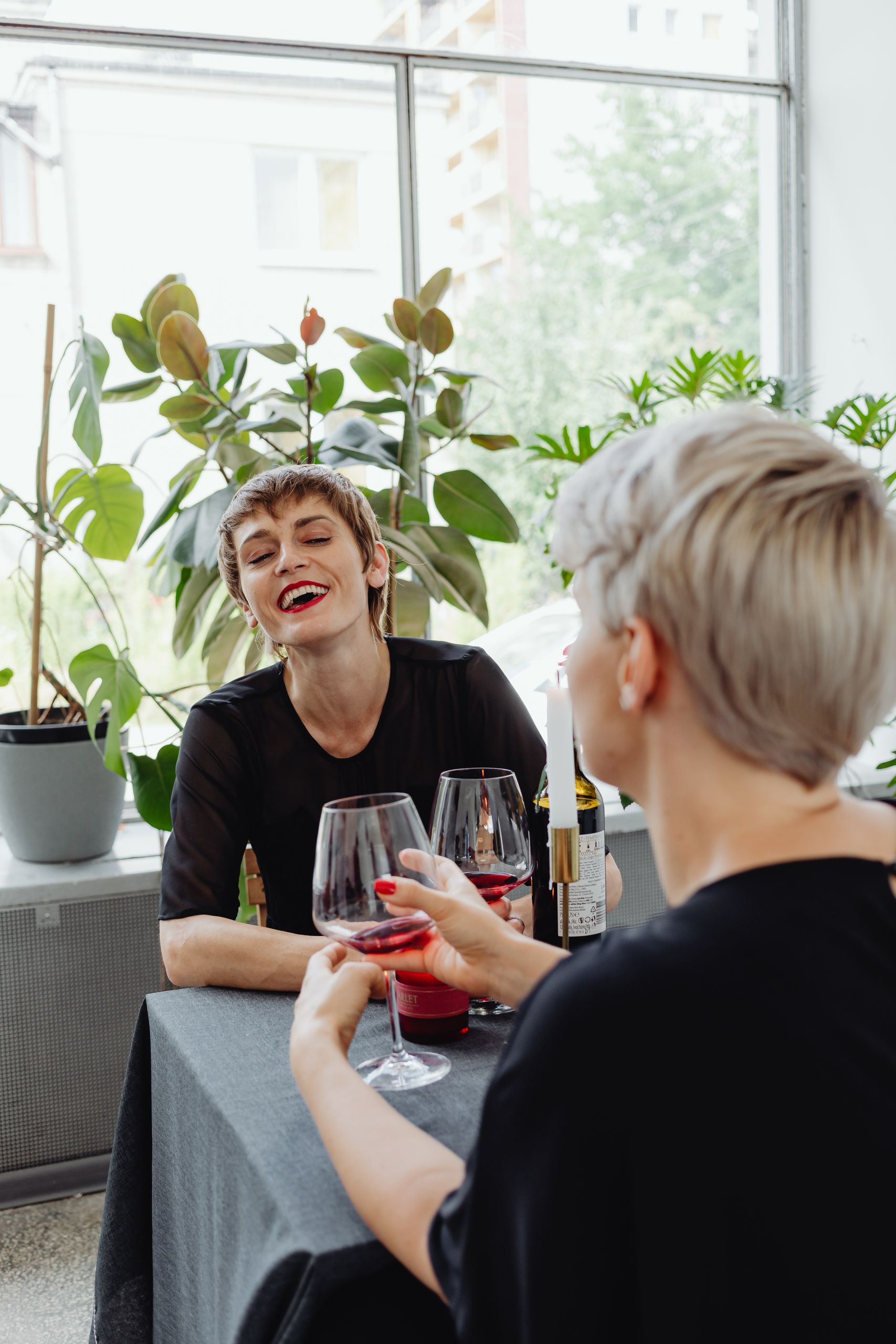 Why I Am Paying Attention to the Stories Women Tell Over Lunch - Two women laughing over glasses of red wine at a stylish indoor restaurant.