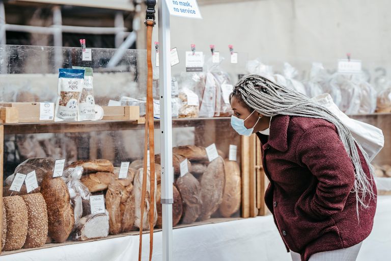 Why I Keep Looking for Local Bread - Side view of adult black lady wearing protective mask and warm coat selecting baking goods while standing in street market in daytime